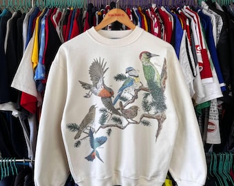May include: Cream-colored sweatshirt featuring a detailed illustration of various birds perched on a tree branch. The birds are depicted in shades of brown, blue, green, and red. The sweatshirt has a crew neck and long sleeves.