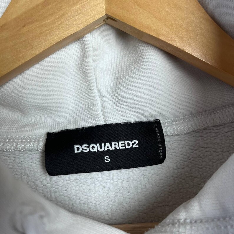 May include: Close-up of a white hoodie with a black label. The label reads "DSQUARED2" in white capital letters, with the letter "S" below. The hoodie is hanging on a wooden hanger.