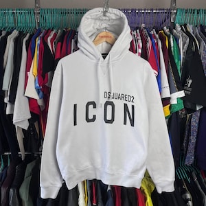May include: White hoodie with a drawstring hood and the text "DSQUARED2 ICON" printed in black. The hoodie is hanging on a wooden hanger against a backdrop of various clothing items.