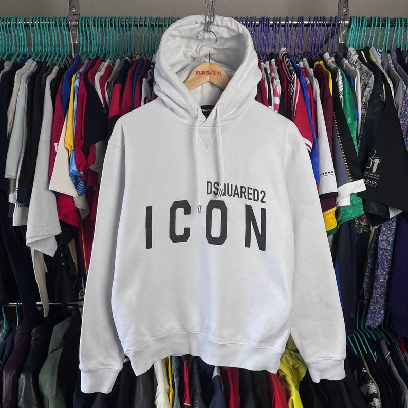 May include: White hooded sweatshirt featuring the text "DSQUARED2 ICON" in black. The hoodie has a drawstring and is displayed on a wooden hanger. A variety of clothes hang in the background.