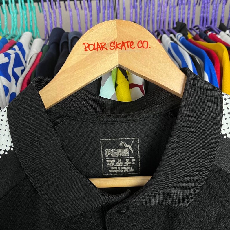 May include: Black polo shirt with a collar and buttoned placket, hanging on a wooden hanger. The hanger has the text "POLAR SKATE CO." in red. The shirt has white polka dots on the shoulder.
