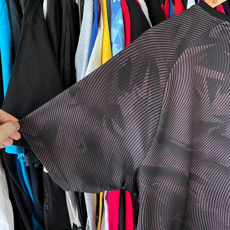 Puede incluir: Una camiseta de manga corta con un patr&oacute;n abstracto en gris oscuro y negro compuesto por finas l&iacute;neas diagonales y curvas. La tela tiene un brillo sutil y una apariencia texturizada, con un cuello redondo negro s&oacute;lido y mangas ragl&aacute;n.