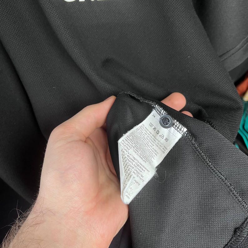 May include: Black polo shirt with a buttoned collar. The shirt has a tag with care instructions and a small button. The fabric appears to be a textured knit.