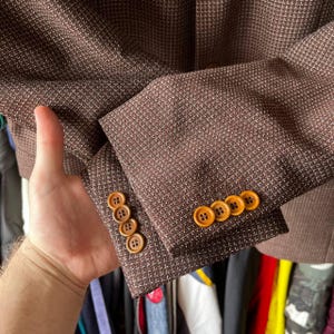 May include: Close-up of a brown blazer with a subtle white grid pattern. The blazer features four brown buttons on the sleeve and three on the cuff. The fabric has a textured appearance, and the buttons have a marbled effect.