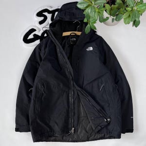 Può includere: Una giacca nera con cappuccio, chiusura a zip e diverse tasche. La giacca ha un logo sul petto sinistro che recita "The North Face".