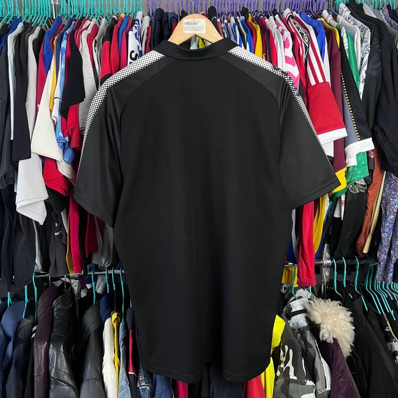 May include: Black polo shirt with white and black checkered accents on the shoulders. The shirt has short sleeves and a collared neckline. The shirt is hanging on a wooden hanger.