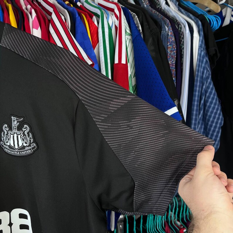 Puede incluir: Una camiseta de f&uacute;tbol negra con el escudo del Newcastle United en el pecho. La manga tiene un patr&oacute;n texturizado gris oscuro con l&iacute;neas onduladas horizontales. La tela es lisa y el escudo es un parche bordado en blanco y negro.