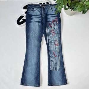 Può includere: Jeans in denim blu con gamba a campana e un designo argento e rosso sulla gamba destra. Il design include la parola "Miss" in un font stilizzato.