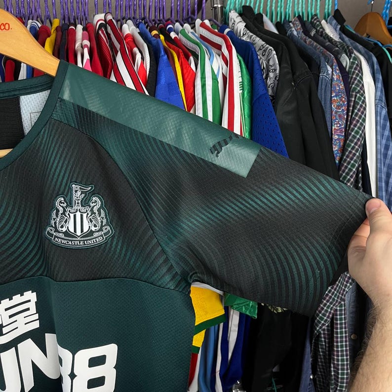 K&ouml;nnte beinhalten: Ein dunkelgr&uuml;nes und schwarzes kurz&auml;rmeliges Sporttrikot mit dem Newcastle United Wappen auf der Brust und einem gro&szlig;en wei&szlig;en Sponsorenlogo. Der Stoff weist ein dezentes wellenf&ouml;rmiges Texturmuster an Schultern und &Auml;rmeln mit schwarzem Logo auf.