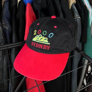 Könnte beinhalten: Schwarze Baseballkappe mit rotem Schirm. Auf der Kappe ist der Text "2000 SYDNEY" in Gelb, Blau und Rot gestickt. Oben befindet sich ein roter Knopf.