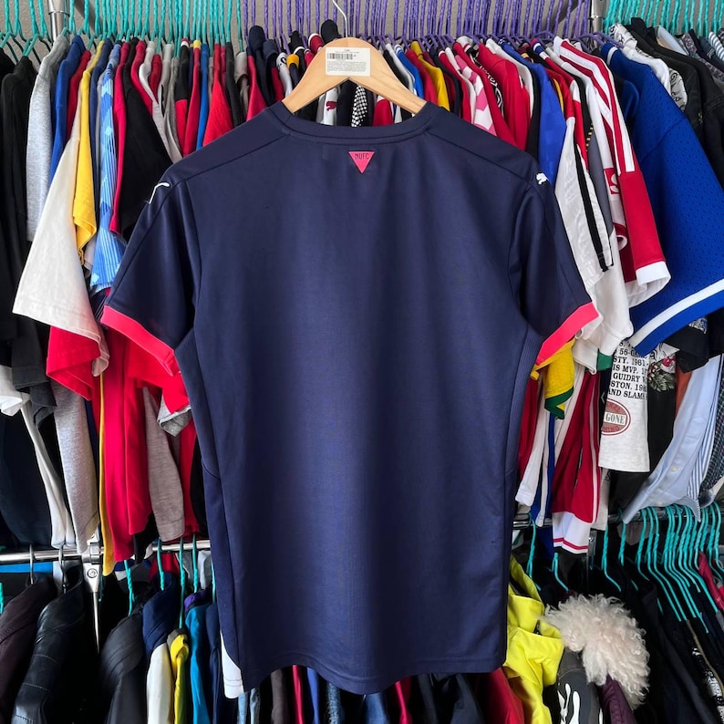 Pu&ograve; includere: Maglietta sportiva blu navy con finiture rosse sulle maniche e un piccolo stemma rosso sul retro. La maglietta &egrave; appesa a una gruccia di legno tra altri capi d'abbigliamento.