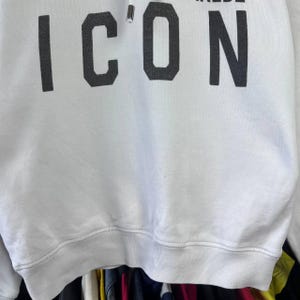 May include: White hooded sweatshirt featuring the word "ICON" in large, dark grey block letters. The sweatshirt has a drawstring hood and a ribbed hem. The text is centered on the chest.