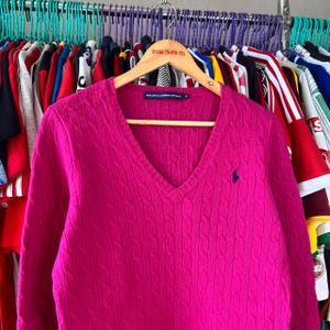 May include: A vibrant pink cable-knit sweater with a deep V-neck. The sweater features a small navy blue logo on the chest. The garment is hanging on a wooden hanger, surrounded by other clothing items on a rack.