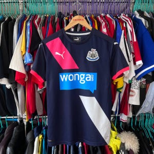 Pu&ograve; includere: Maglia da calcio Puma blu navy del Newcastle United con dettagli rosa e bianchi. Presenta lo stemma del club e la scritta "wonga.com" in blu. &Egrave; visibile anche il logo Puma.