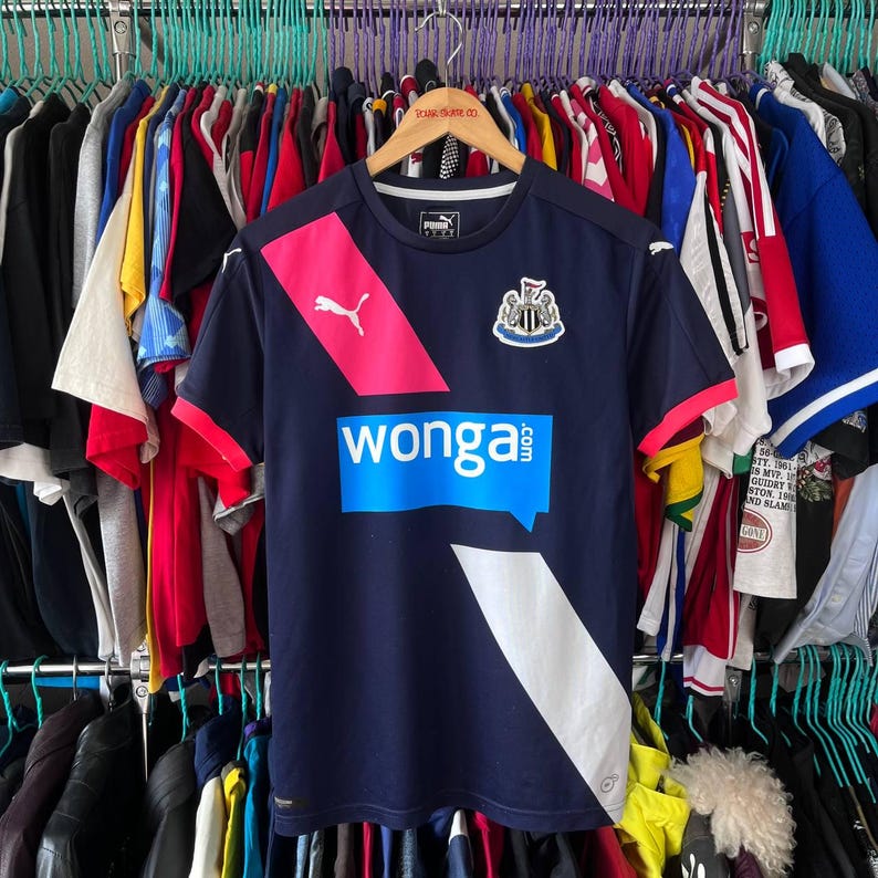 Pu&ograve; includere: Maglia da calcio Puma blu navy del Newcastle United con dettagli rosa e bianchi. Presenta lo stemma del club e la scritta "wonga.com" in blu. &Egrave; visibile anche il logo Puma.