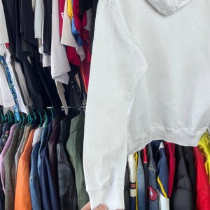 May include: A white hoodie with a drawstring hood and ribbed cuffs. The hoodie is made of a soft material and is hanging on a rail with other clothing items.