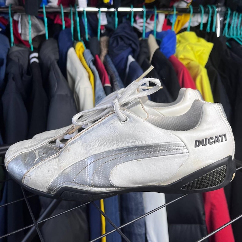 May include: White and silver Puma sneakers with the word "Ducati" printed on the side. The sneakers have black soles and white laces. The Puma logo is visible on the side of the shoe.