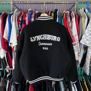 Könnte beinhalten: Schwarze Bomberjacke mit weißen Streifen und dem Text "LYNCHBURG Tennessee U.S.A." in Weiß. Die Jacke hängt an einem Kleiderständer mit anderen Kleidungsstücken.