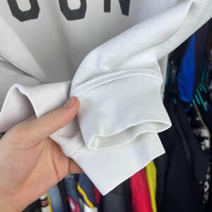 May include: Close-up of a white sweatshirt sleeve with a ribbed cuff. The fabric appears to be a soft, medium-weight material. The cuff is a solid white colour, and the stitching is visible.