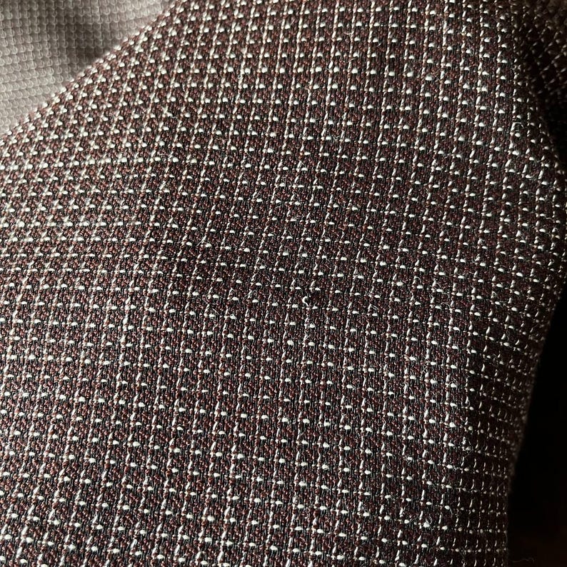 May include: Close-up of a textured fabric with a repeating pattern of small white dots on a dark brown background. The fabric appears to be woven, with a subtle sheen. The overall effect is a sophisticated, geometric design.