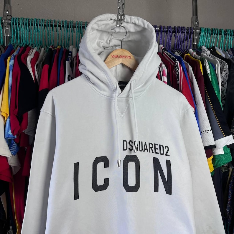 May include: White hooded sweatshirt featuring the text "DSQUARED2 ICON" in black. The sweatshirt has a drawstring hood and a front pocket. The garment is displayed on a wooden hanger.