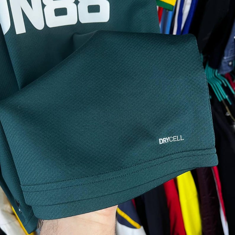 K&ouml;nnte beinhalten: Ein dunkelgr&uuml;nes Sporttrikot mit einem wei&szlig;en Grafikdruck im oberen Bereich und dem Schriftzug "DRYCELL" in Wei&szlig; nahe dem unteren Saum. Der Stoff hat ein dezentes Texturmuster und einen geraden Saum mit durchgehenden Doppelsteppn&auml;hten.