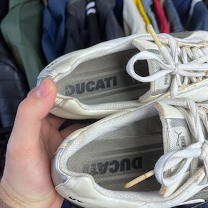 May include: Pair of white and silver sneakers with the word "DUCATI" printed on the insoles. The sneakers have white laces and a silver accent on the side. The Puma logo is visible on the side of the shoe.
