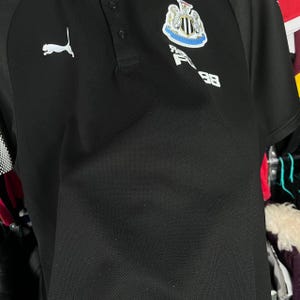 May include: Black polo shirt with a white Puma logo on the right shoulder. The shirt features a Newcastle United crest and the number 98. The shirt has a collared neckline and short sleeves.