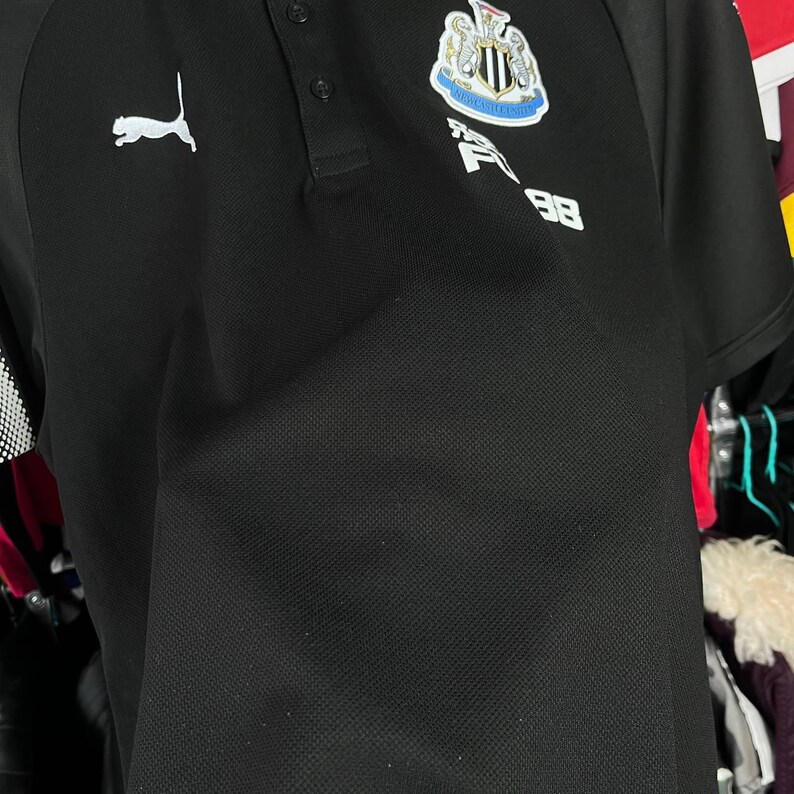 May include: Black polo shirt with a white Puma logo on the right shoulder. The shirt features a Newcastle United crest and the number 98. The shirt has a collared neckline and short sleeves.