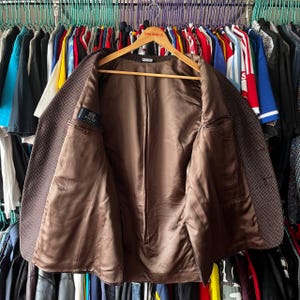 May include: A brown blazer with a subtle checkered pattern, displayed open on a wooden hanger. The interior lining is a solid brown, and a small label is visible on the inside. The blazer is surrounded by a variety of clothing items.