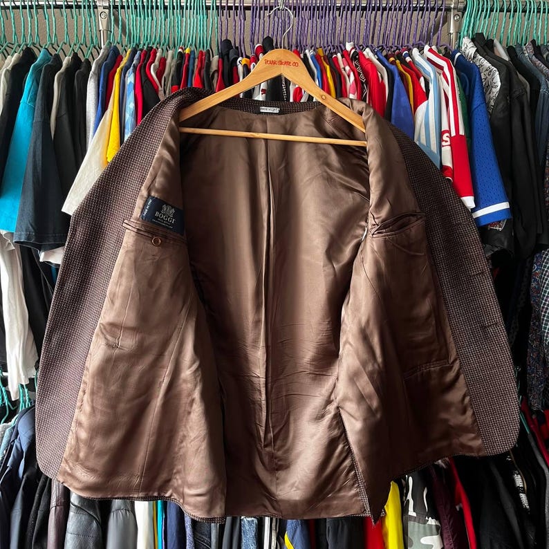May include: A brown blazer with a subtle checkered pattern, displayed open on a wooden hanger. The interior lining is a solid brown, and a small label is visible on the inside. The blazer is surrounded by a variety of clothing items.
