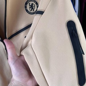 Puede incluir: Chaqueta con cremallera beige con detalles negros y un parche con el logotipo del Chelsea Football Club. La chaqueta tiene un bolsillo con cremallera negro y una raya negra. El logotipo es circular, con un emblema floral y las palabras "Chelsea Football Club".