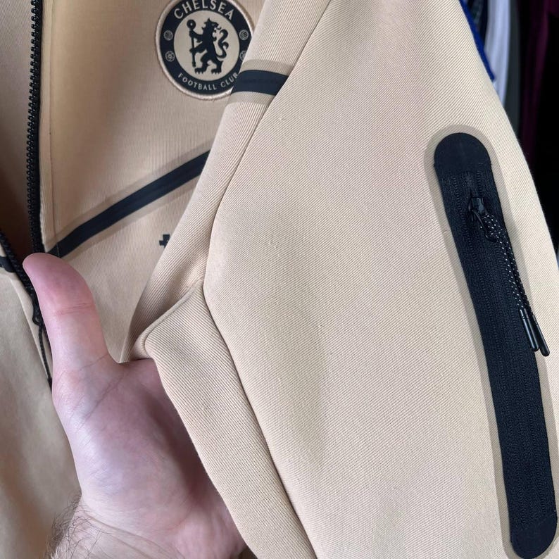 Puede incluir: Chaqueta con cremallera beige con detalles negros y un parche con el logotipo del Chelsea Football Club. La chaqueta tiene un bolsillo con cremallera negro y una raya negra. El logotipo es circular, con un emblema floral y las palabras "Chelsea Football Club".