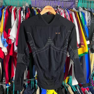 Puede incluir: Camiseta deportiva negra de manga larga con cuello en V. La camiseta presenta un diseño texturizado en los hombros y las mangas, con costuras en contraste. Un logotipo naranja es visible en el pecho. La camiseta se exhibe en una percha con otras prendas.