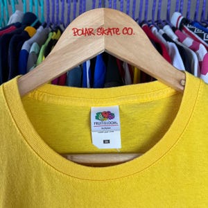 Puede incluir: Una camiseta amarilla brillante con cuello redondo, colgada de una percha de madera. La percha muestra el texto rojo "POLAR SKATE CO.". La camiseta tiene una etiqueta "FRUIT OF THE LOOM".