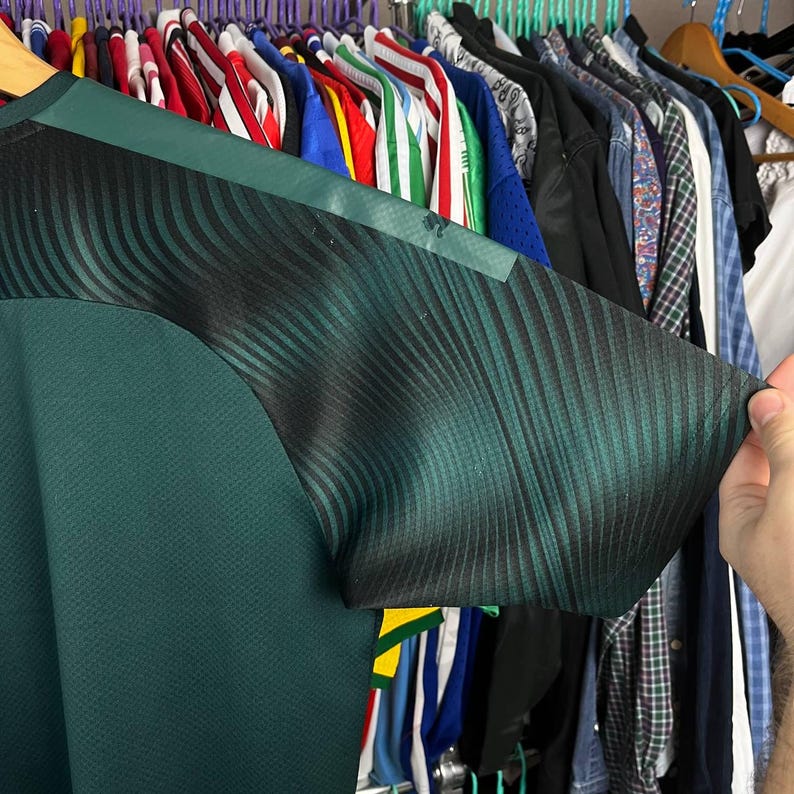 K&ouml;nnte beinhalten: Ein dunkelgr&uuml;nes Sporttrikot mit einem strukturierten, einfarbig gr&uuml;nen Rumpf und kurzen &Auml;rmeln, die ein schwarz-dunkelgr&uuml;nes Verlaufsmuster aus feinen, diagonalen Wellenlinien aufweisen, das sich &uuml;ber die Schulter bis zum &Auml;rmel erstreckt.