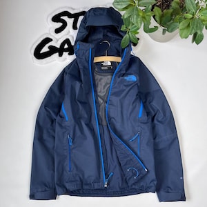 Könnte beinhalten: Eine marineblaue Kapuzenjacke mit blauen Besätzen und einem Reißverschluss. Die Jacke hat ein Logo auf der linken Brust, das "The North Face" lautet.