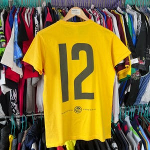 Puede incluir: Una camiseta amarilla con el n&uacute;mero "12" impreso en gris oscuro en la espalda. La camiseta tiene un peque&ntilde;o gr&aacute;fico y texto en la parte inferior. La camiseta est&aacute; colgada en una percha con otras prendas.