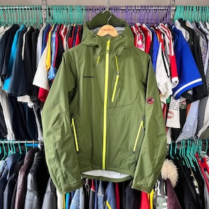 May include: Olive green hooded jacket with bright yellow zippers and accents. The jacket features the brand name "Mammut" on the chest and a logo on the sleeve. It has multiple zippered pockets and a full-length front zipper. The jacket is hanging on a rack with other clothing.