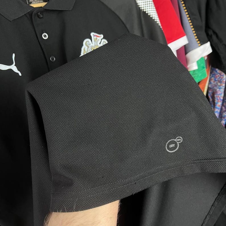 May include: Black polo shirt with a white Puma logo and a team crest. The shirt has a textured fabric and a Dry Cell logo. The shirt has a three-button placket and a collar.