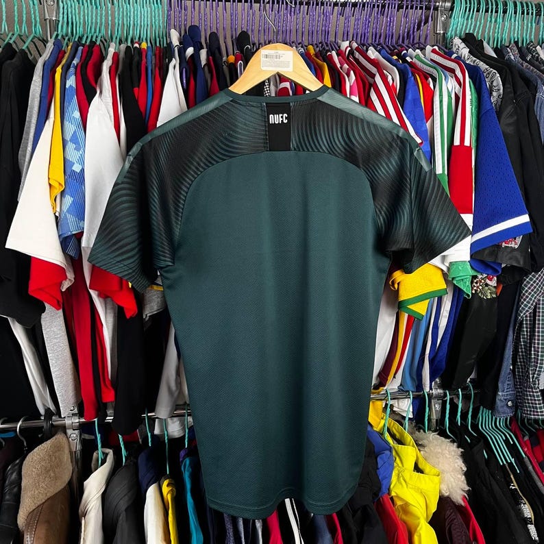 K&ouml;nnte beinhalten: Ein dunkelgr&uuml;nes kurz&auml;rmeliges Sporttrikot mit schwarzen Raglan&auml;rmeln, die ein dezentes Wellenmuster aufweisen. Im Nackenbereich befindet sich ein schwarzer rechteckiger Aufn&auml;her mit dem wei&szlig;en Text NUFC in einer serifenlosen Schrift.
