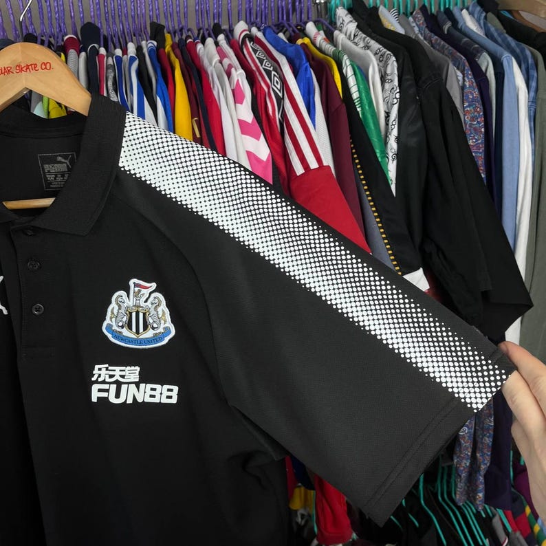 May include: Black Puma polo shirt with white dotted accents on the sleeves. Features the Newcastle United crest and the Fun88 logo. The shirt is hanging on a wooden hanger.