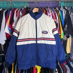 May include: A vintage Rothmans racing jacket in cream, navy blue, and red. The jacket features a zip-up front, a stand-up collar, and the Rothmans logo on the chest. The jacket has horizontal red stripes across the chest and sleeves.