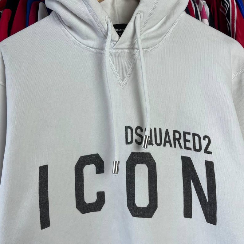 May include: A white hooded sweatshirt featuring the text "DSQUARED2 ICON" in black. It has a drawstring hood, a kangaroo pocket, and ribbed cuffs and hem. The material looks to be a soft, medium-weight fabric.
