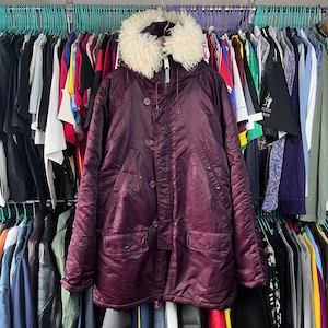 Könnte beinhalten: Ein burgunderfarbener Wintermantel mit einer mit Kunstfell gefütterten Kapuze. Der Mantel hat eine Knopfleiste, zwei große Vordertaschen und einen Reißverschluss. Der Mantel hängt an einem Kleiderständer mit anderen Kleidungsstücken.