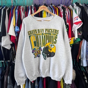 Puede incluir: Sudadera gris claro de los Green Bay Packers con el texto "Green Bay Packers 1996 Super Bowl Champions". El diseño incluye un casco de fútbol y el logotipo del Super Bowl XXXI en verde, amarillo y morado.