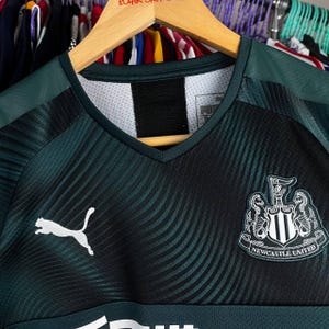 K&ouml;nnte beinhalten: Ein dunkelgr&uuml;nes und schwarzes Fu&szlig;balltrikot mit V-Ausschnitt, einem wei&szlig;en Puma-Logo auf der rechten Brust und dem Newcastle United-Wappen auf der linken Brust. Der Stoff hat ein dezentes Wellenmuster auf der Vorderseite und einfarbige Schultern.