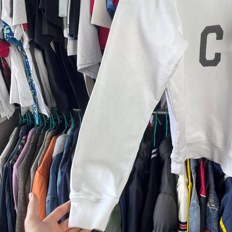 May include: A white sweatshirt featuring a large, dark grey letter 'C'. The long sleeve is visible, with a ribbed cuff. The sweatshirt is hanging amongst other clothing items on hangers.