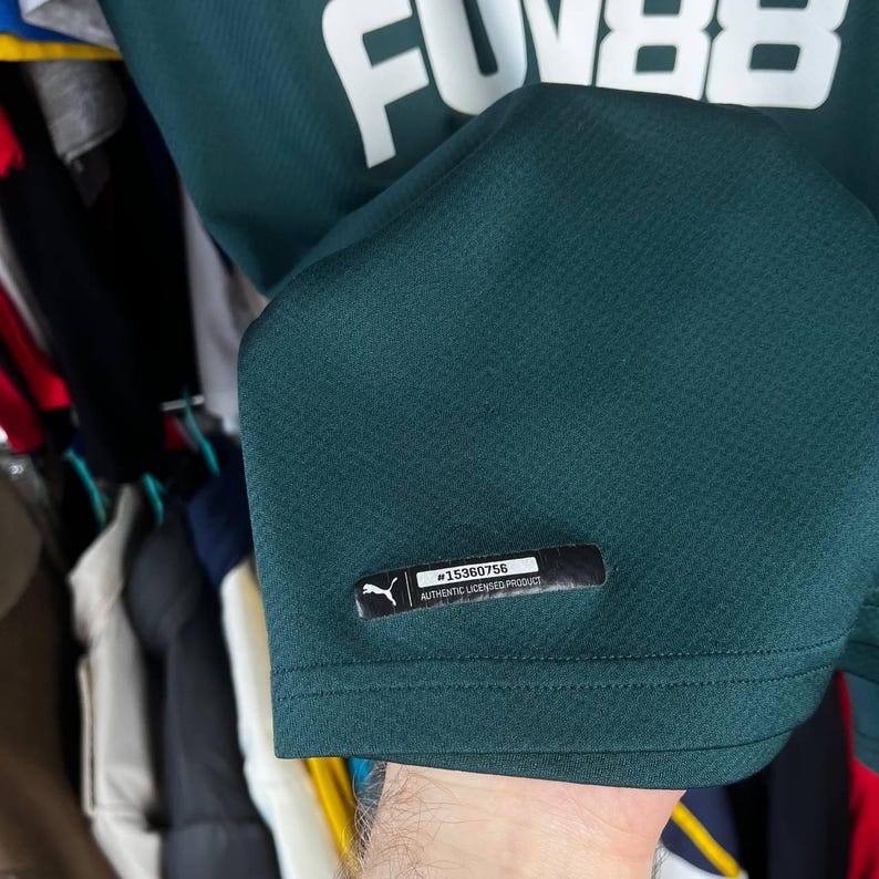 K&ouml;nnte beinhalten: Ein dunkelgr&uuml;nes Sporttrikot mit strukturierter Stoffoberfl&auml;che. Im oberen Bereich ist der wei&szlig;e Schriftzug FUN88 zu sehen. Nahe dem unteren Saum befindet sich ein schwarzes rechteckiges Etikett mit dem Puma-Logo und dem Text #15360756 AUTHENTIC LICENSED PRODUCT.