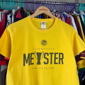 Puede incluir: Una camiseta amarilla con cuello redondo y mangas cortas. La camiseta presenta el texto "SCHWEIZER MEYSTER 2018" en negro, junto con un logotipo circular. La camiseta est&aacute; colgada en una percha de madera entre otras prendas.